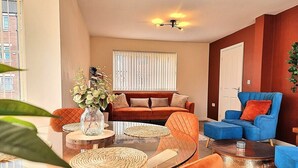 A modern living room with a glass coffee table, orange and blue sofas, and a dining area with wicker placemats. - Detached House w/Free Parking | Long Stay Discount (Leicestershire)