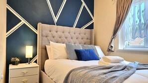 A bedroom with a tufted headboard, a bedside table with a lamp, and a window with curtains. - Detached House w/Free Parking | Long Stay Discount (Leicestershire)