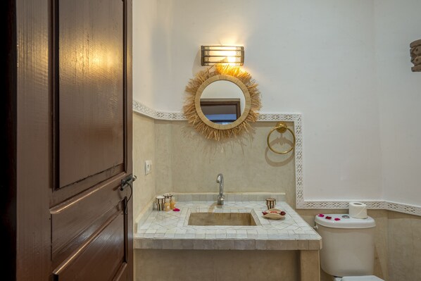 Un baño con puerta de madera, un lavamanos con espejo, un inodoro y una lámpara decorativa en la pared.