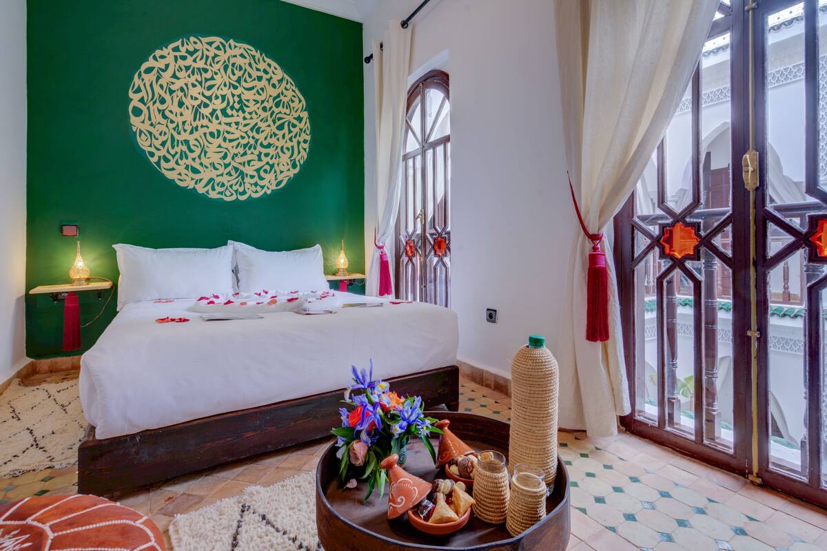 Un lit bien fait, avec du linge de lit blanc, un élément décoratif mural, une table de chevet avec une lampe, et une vue sur l’extérieur par une fenêtre agrémentée d’une porte décorative.