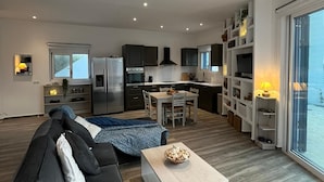 Ein modernes Wohnzimmer mit einer schwarzen Couch, einem Couchtisch und einem Essbereich mit Tisch und Stühlen. Im Hintergrund ist eine Küche mit dunklen Schränken und einem Fenster zu sehen.