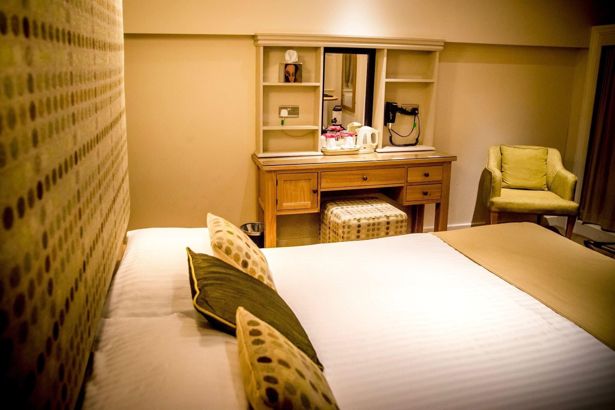 Une chambre d’hôtel comprenant un lit, une commode en bois avec un miroir, une chaise et une petite table avec une bouilloire et des tasses.
