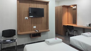 Habitación de hotel con televisión instalada en un panel de madera, un escritorio con silla, dos camas con toallas y un espejo.