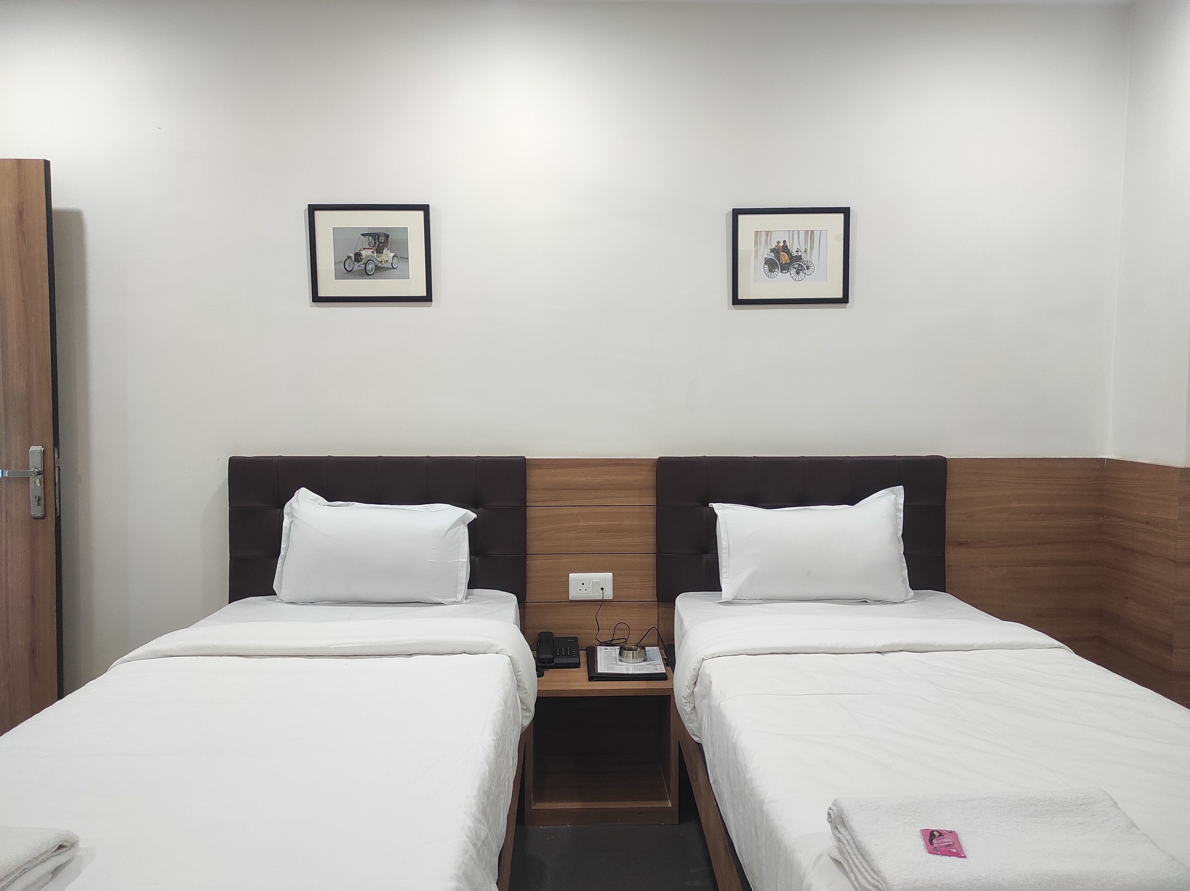 Habitación de hotel con dos camas individuales, cabecera de madera, mesita de noche con teléfono y lámpara, y dos cuadros enmarcados en la pared.