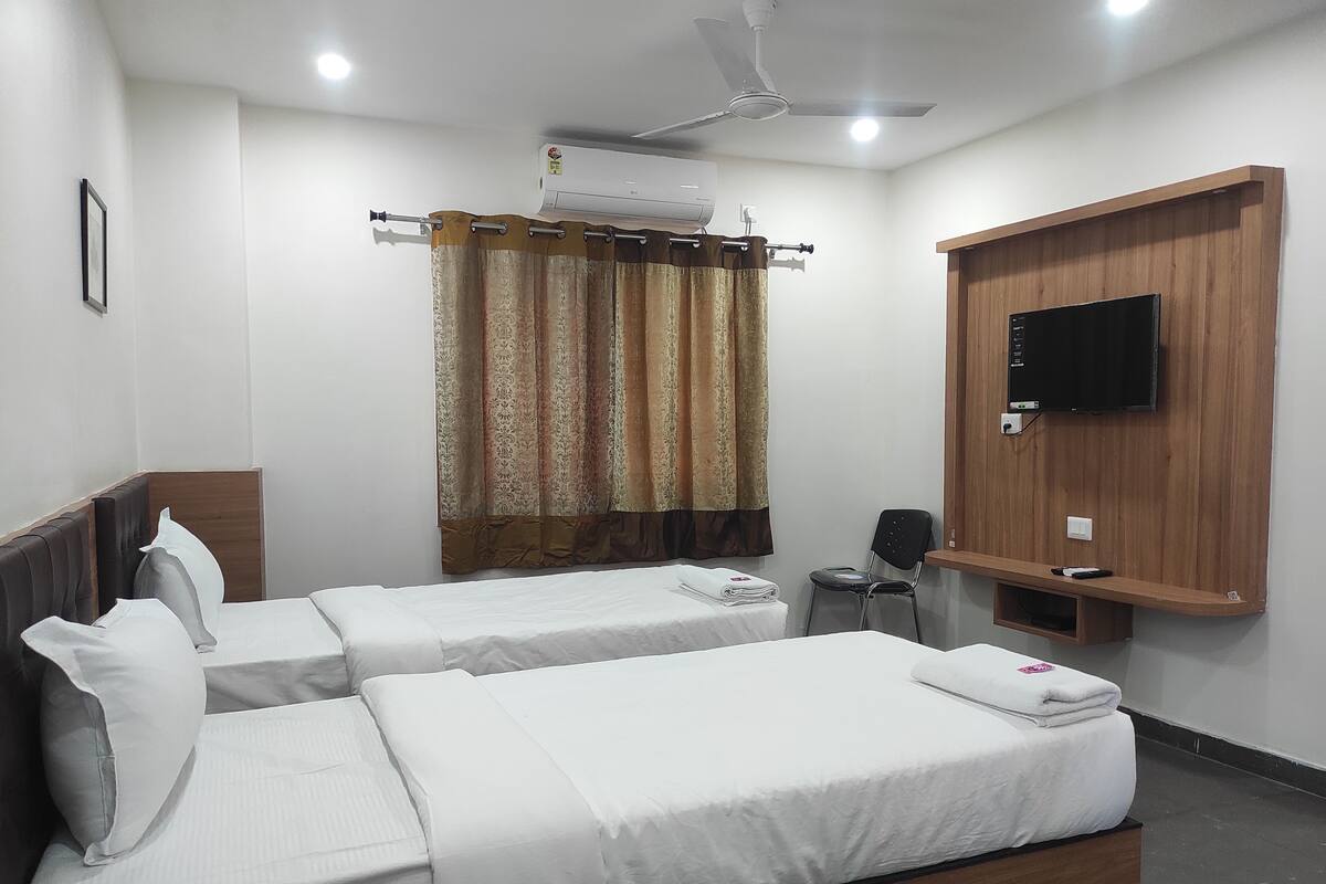 A hotel room with two single beds, a ceiling fan, an air conditioning unit, a TV mounted on a wooden panel, and a chair.