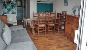 A living room with a grey sofa, a dining table with chairs, a kitchen area with white cabinets, and a tiled floor. - Grand Studio à Quelques pas de la Plage et des Activities de Loisirs (GUIDEL)