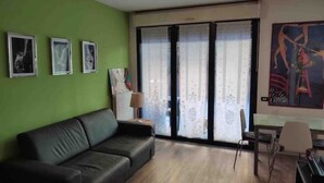 A living room with a black leather sofa, a dining table with chairs, a piano, and a large window with sheer curtains. - La Cà del Lori (Milano)