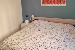 A bedroom with a bed covered in alphabet-patterned bedding, a nightstand with a lamp, and a wall with a red abstract painting.