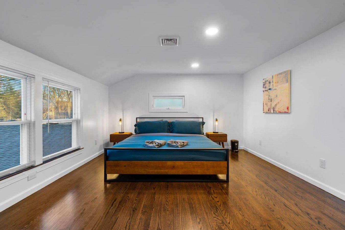 A bedroom with a bed, two bedside tables, a window with blinds, a ceiling vent, and a wall-mounted artwork.