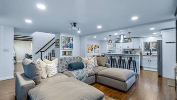 A modern living room with a sectional sofa, a kitchen in the background, and a staircase leading to another level.