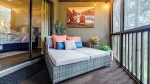 A cozy balcony with a wicker sofa bed, a painting of chairs by a lake, and a view of trees. - Pet Friendly Condo with Screened Deck & Fireplace (Jasper)