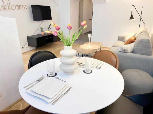 Un espace de salle à manger moderne avec une table ronde blanche dressée pour quatre personnes, agrémenté d’un vase de tulipes, d’un téléviseur à écran plat fixé au mur et d’un canapé en arrière-plan.