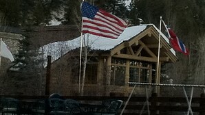 A wooden cabin with a snow-covered roof, an American flag, and a British flag.