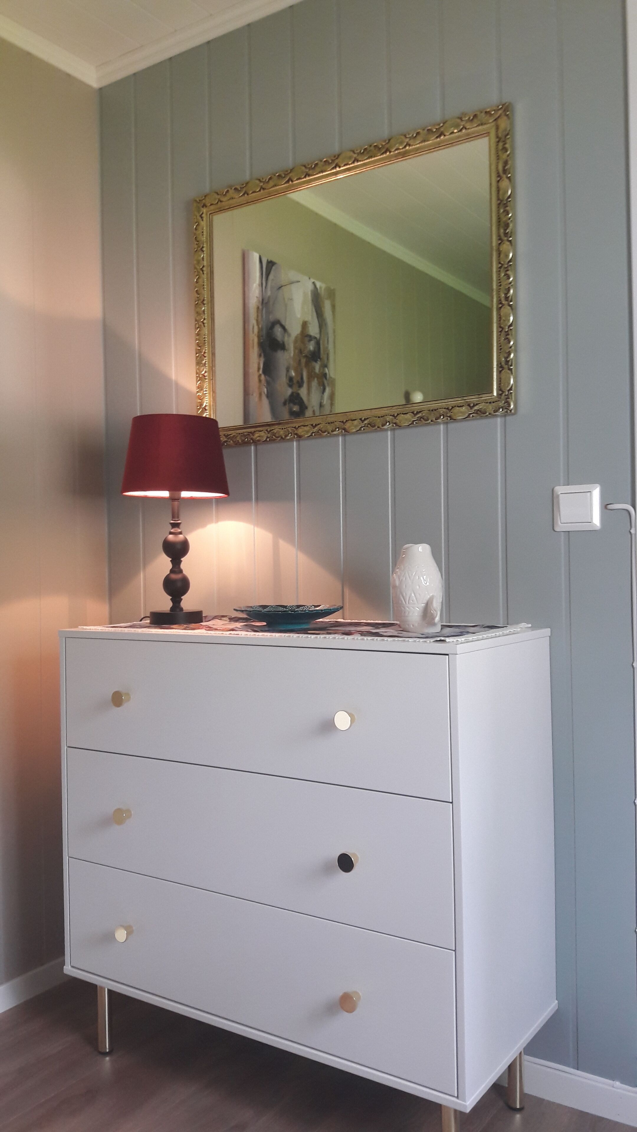 A white dresser with three drawers, a red lamp, a decorative bowl, and a white vase.