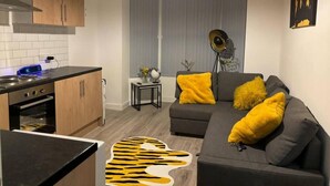 A modern apartment with a kitchen, a grey sofa with yellow cushions, and a large window with blinds. - Apartment in Leeds Close to City Centre (Leeds)