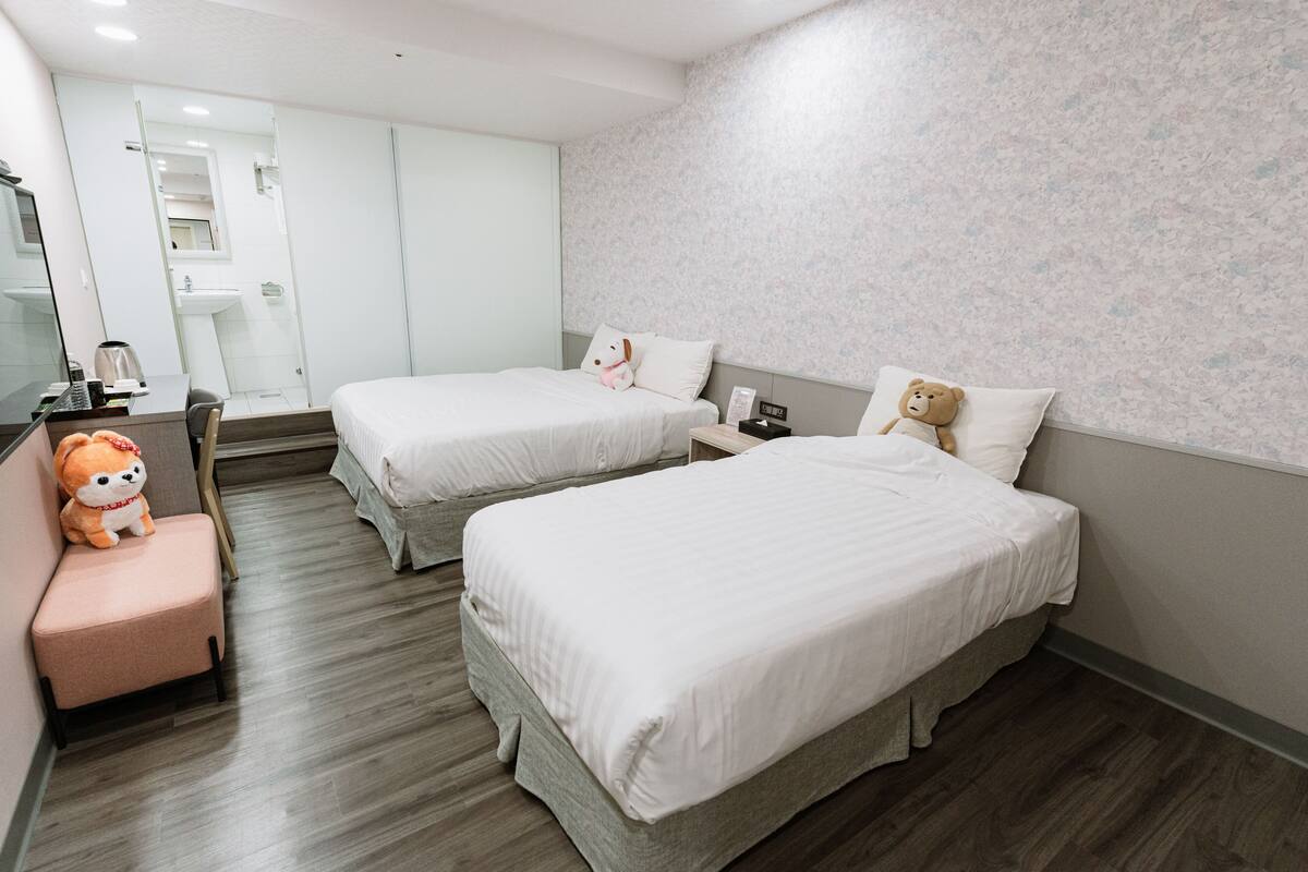 Une chambre d’hôtel avec deux lits, un bureau avec une chaise, un petit canapé, un ours en peluche et un miroir.