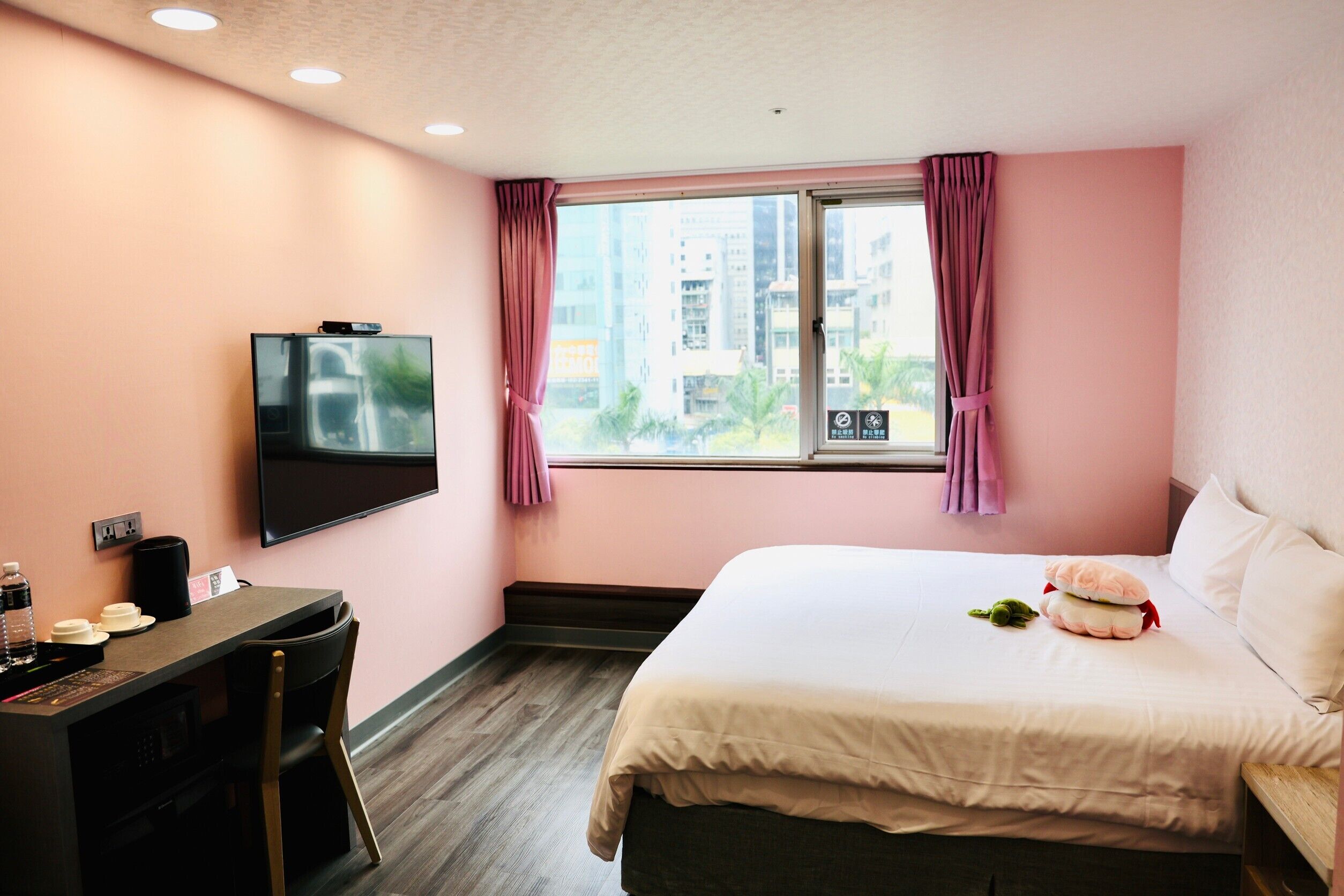 A hotel room with a bed, a desk with a chair, a TV mounted on the wall, and a window with curtains.