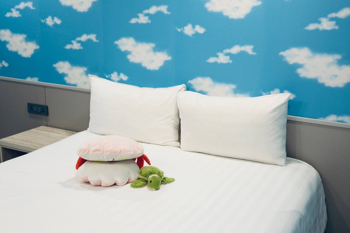 Un lit bien fait, avec une literie blanche, un oreiller orné d’un jouet en peluche, et un mur décoré d’un motif représentant un ciel bleu et des nuages.