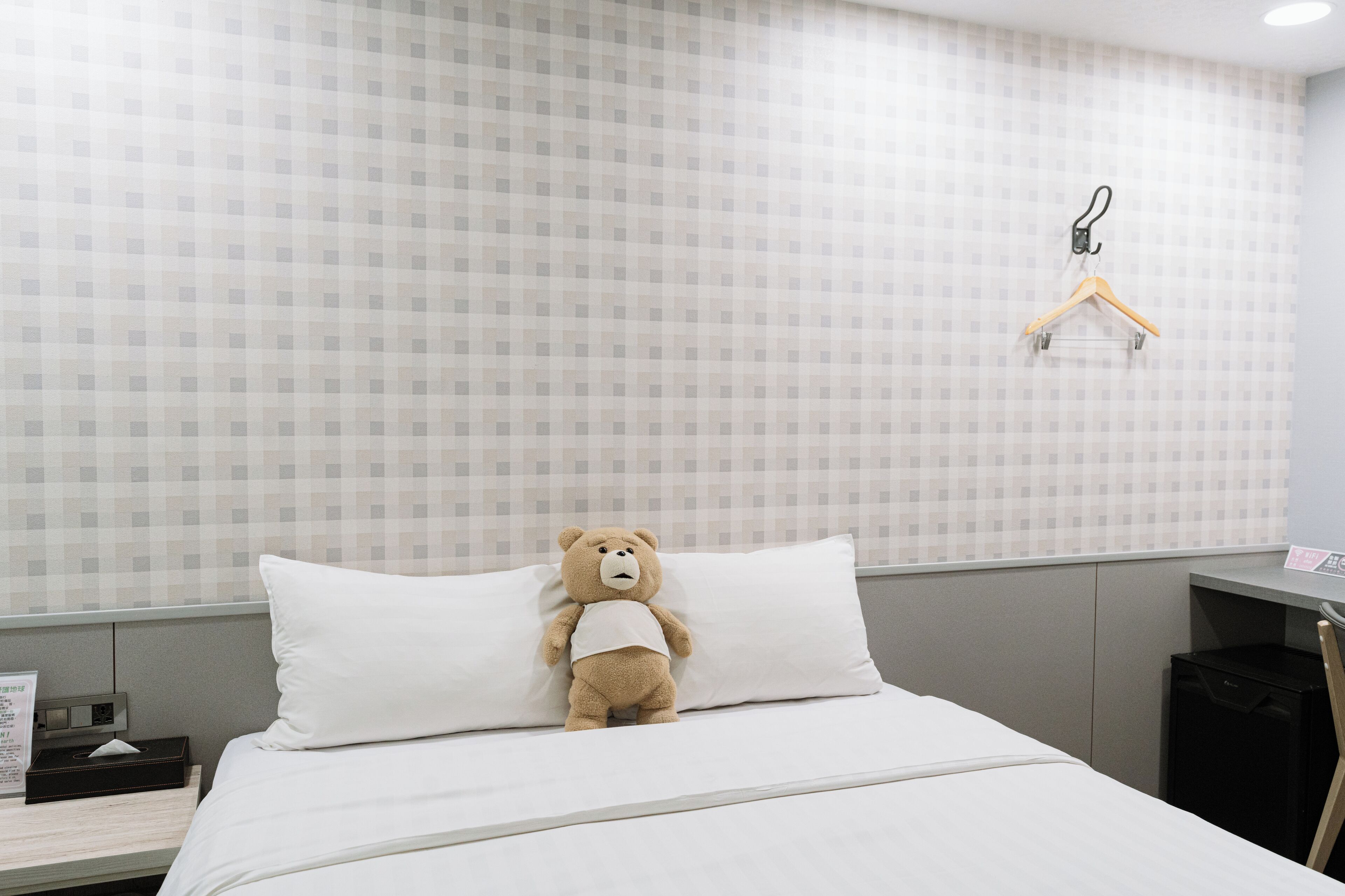 Une chambre d’hôtel avec un lit, un oreiller blanc, un ours en peluche, une petite table avec une boîte de mouchoirs et un porte-manteau fixé au mur.