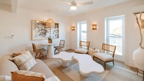 Ein modernes Wohnzimmer mit einem weißen Sofa, zwei weißen Poufs, einem runden Couchtisch und einem Esstisch mit Stühlen. Eine Deckenleuchte, zwei Wandlampen und ein großes Fenster mit Blick auf die Umgebung sind vorhanden.