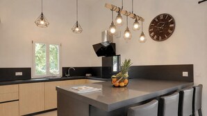 Une cuisine moderne avec un îlot central, des luminaires suspendus, une horloge murale et un panier de fruits.