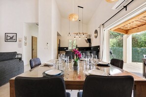 Un espace de repas moderne avec une grande table dressée pour quatre personnes, une cuisine en arrière-plan et une vue sur la verdure à l’extérieur.