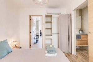 Une chambre à coucher comprenant un lit, une lampe de chevet, une armoire avec des étagères, et une salle de bain avec un lavabo et un miroir.