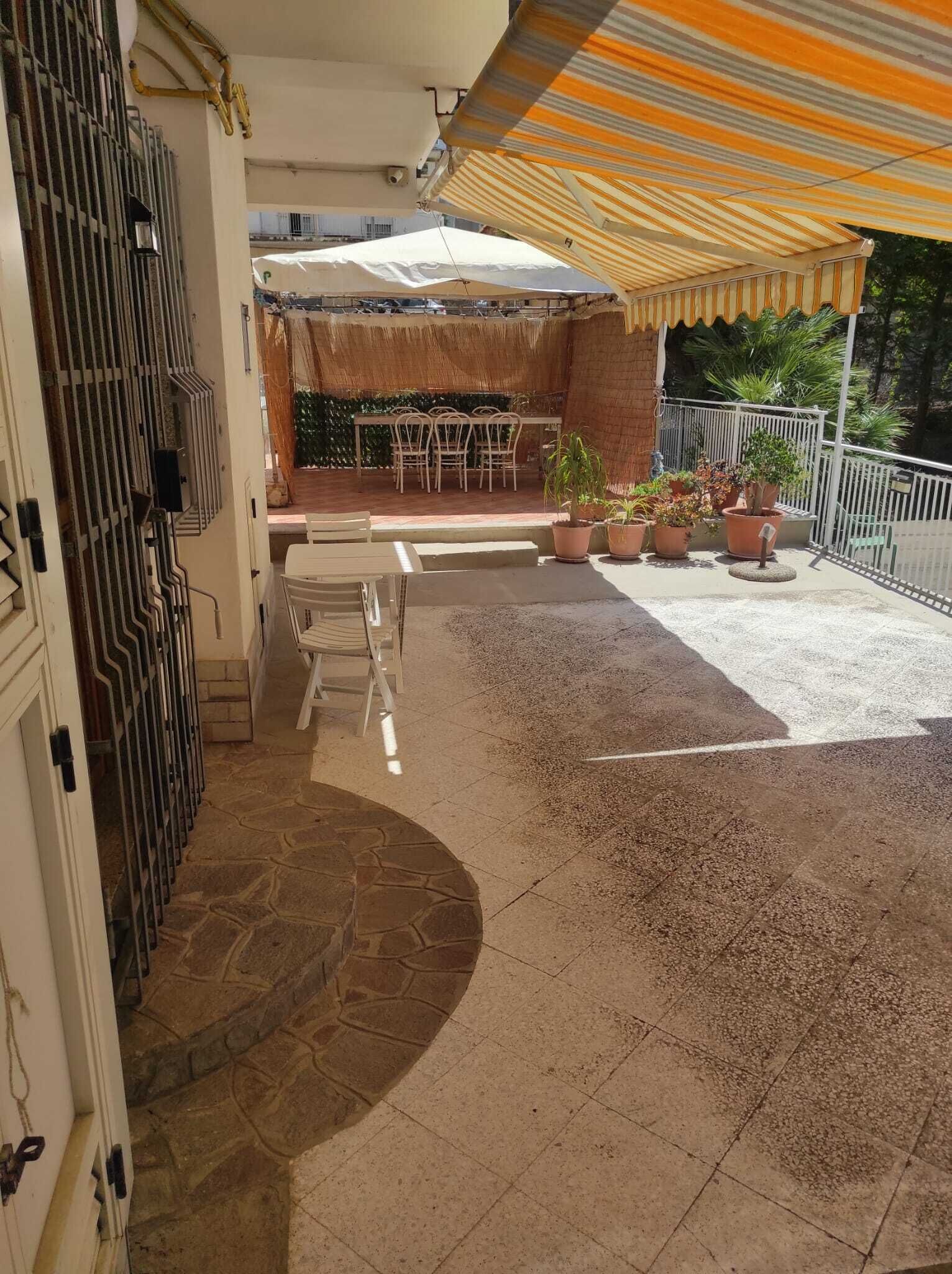 Een terras buiten met een tafel, stoelen, potplanten en een gestreept zonnescherm.