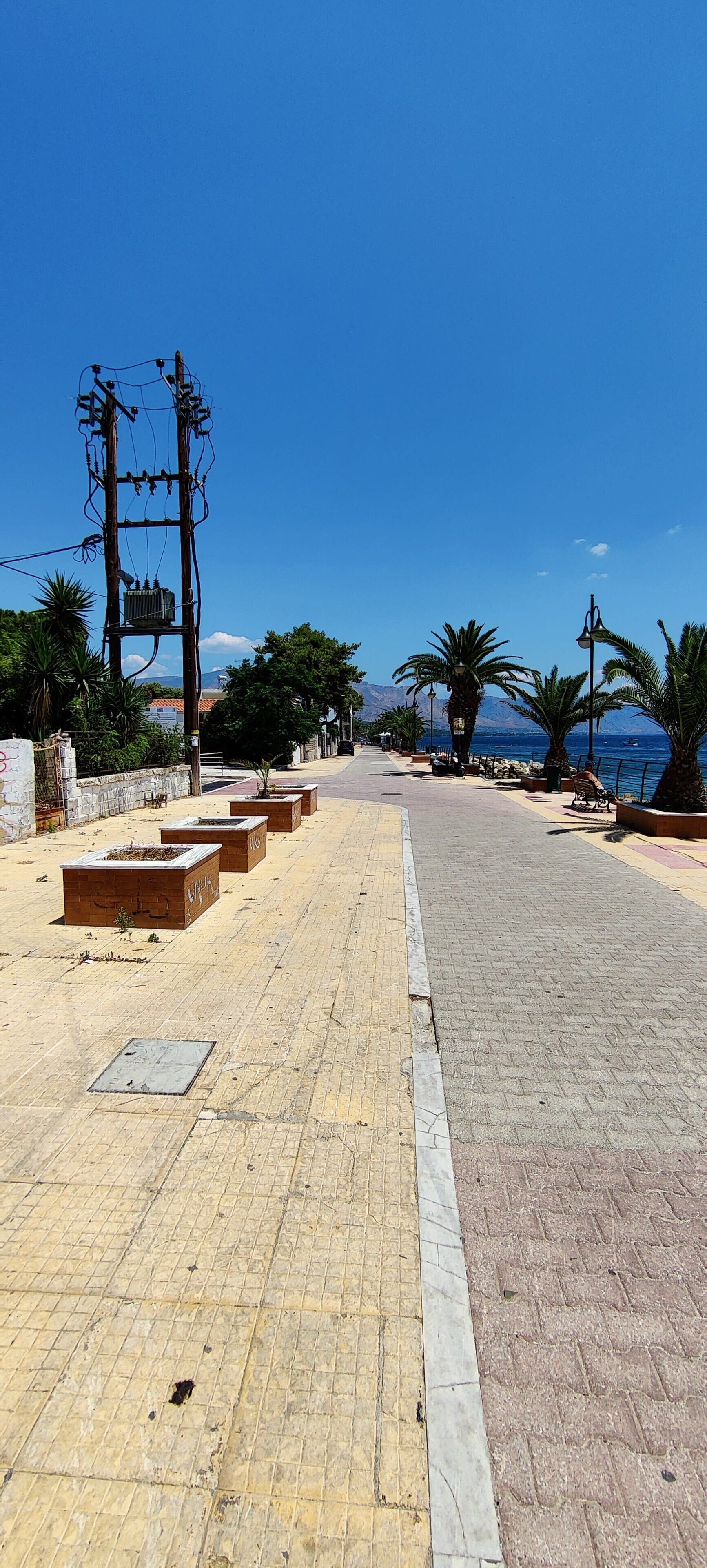 A paved walkway with planters and street lamps, leading to a body of water with mountains in the background.
