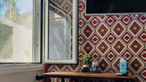 Quarto com parede estampada, janela com persianas, mesa de madeira com planta em vaso e TV fixada na parede.
