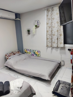 Une chambre à coucher comprenant un lit, un climatiseur fixé au mur, une fenêtre avec des rideaux à motifs, une tête de lit décorative et une petite table de chevet agrémentée d’une plante.