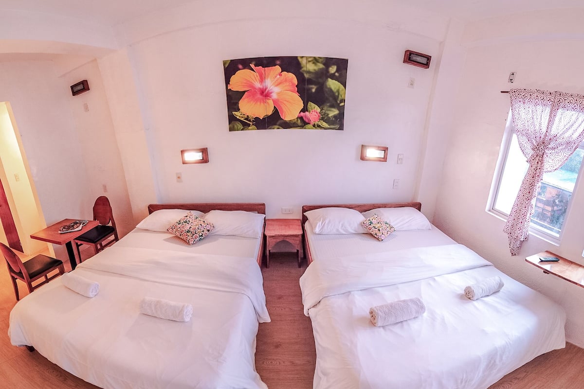A double room with two beds, a desk, and a chair. There is a large floral painting on the wall and a window with a curtain.