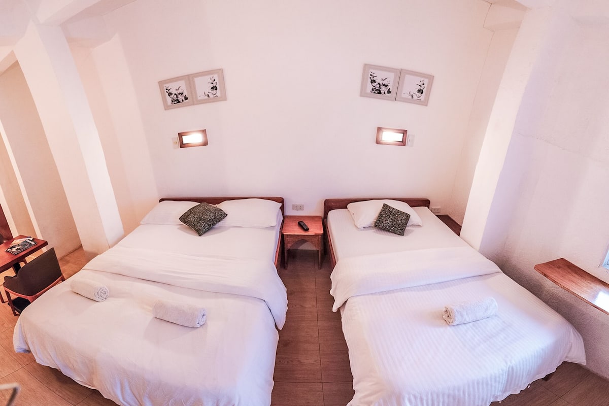 A twin room with two beds, each with a pillow and a blanket, a nightstand with a lamp, and a small table with a magazine.