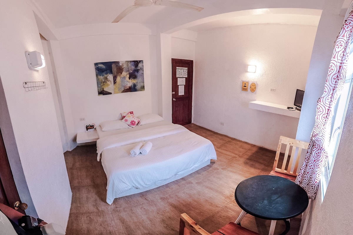 A bedroom with a bed, a desk, a chair, a ceiling fan, and a door.