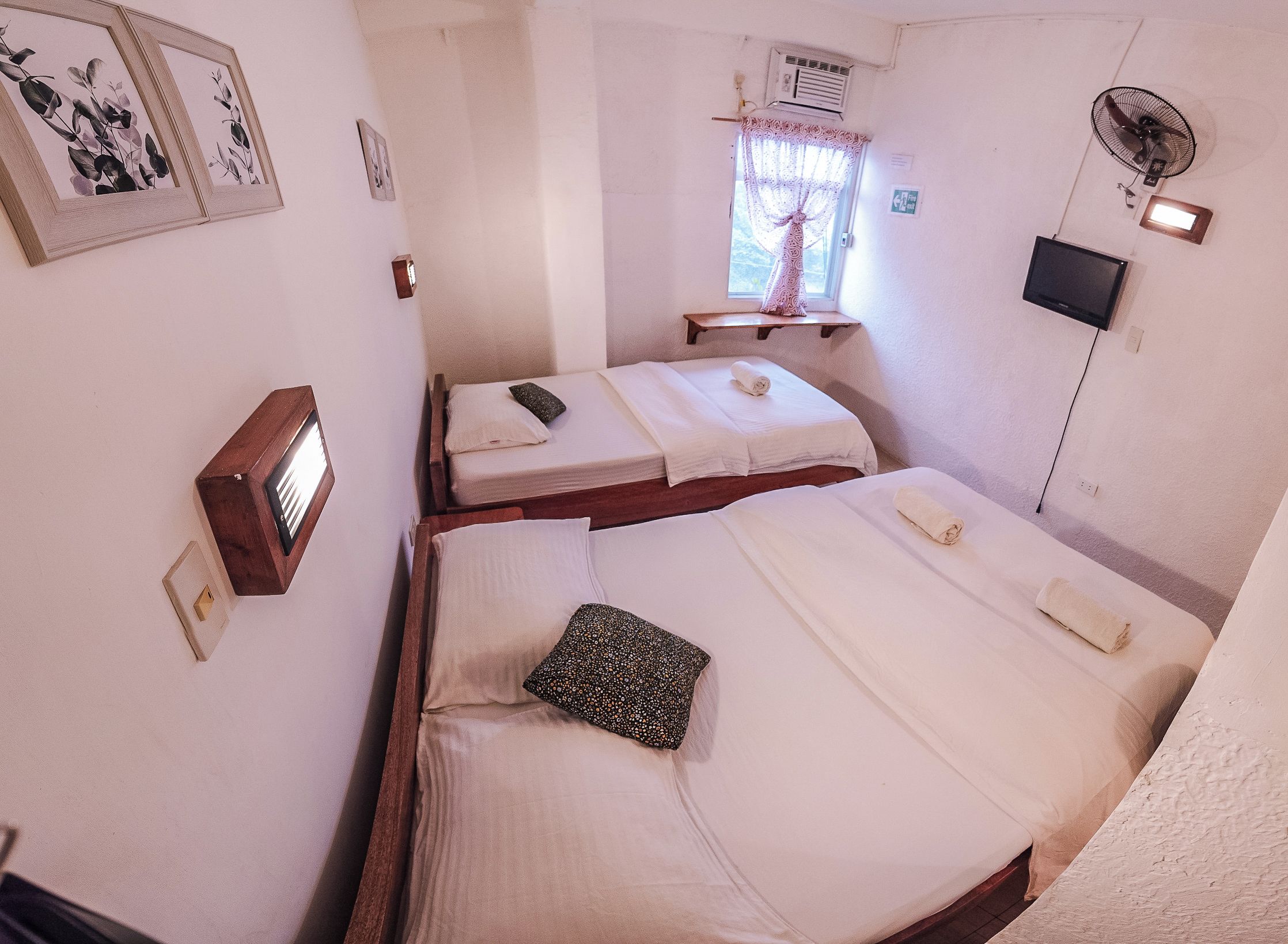 A small, compact hotel room with two beds, a window with lace curtains, a wall-mounted TV, and a ceiling fan.
