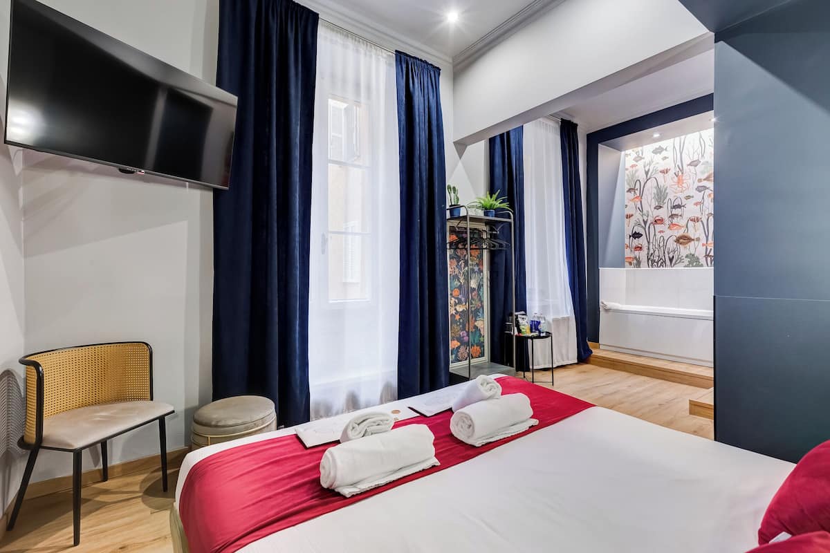 A modern hotel room with a large bed, a flat-screen TV, a wicker chair, a small stool, and a window with curtains.