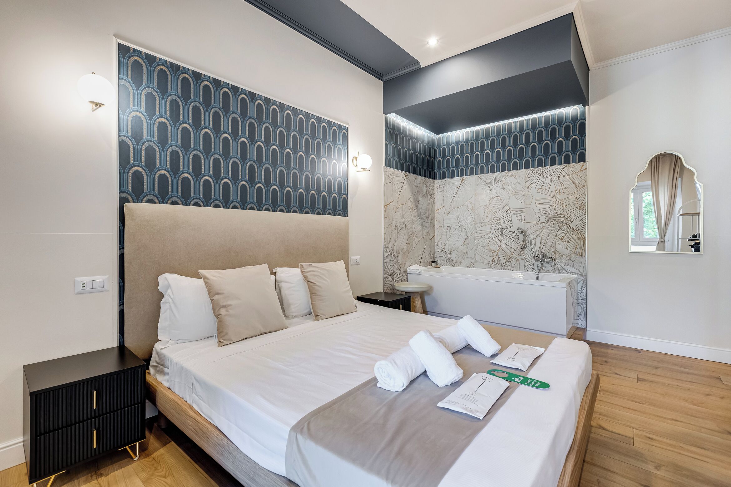 Une chambre d’hôtel moderne avec un grand lit, une petite table de chevet, une baignoire intégrée et un miroir au-dessus du lavabo.