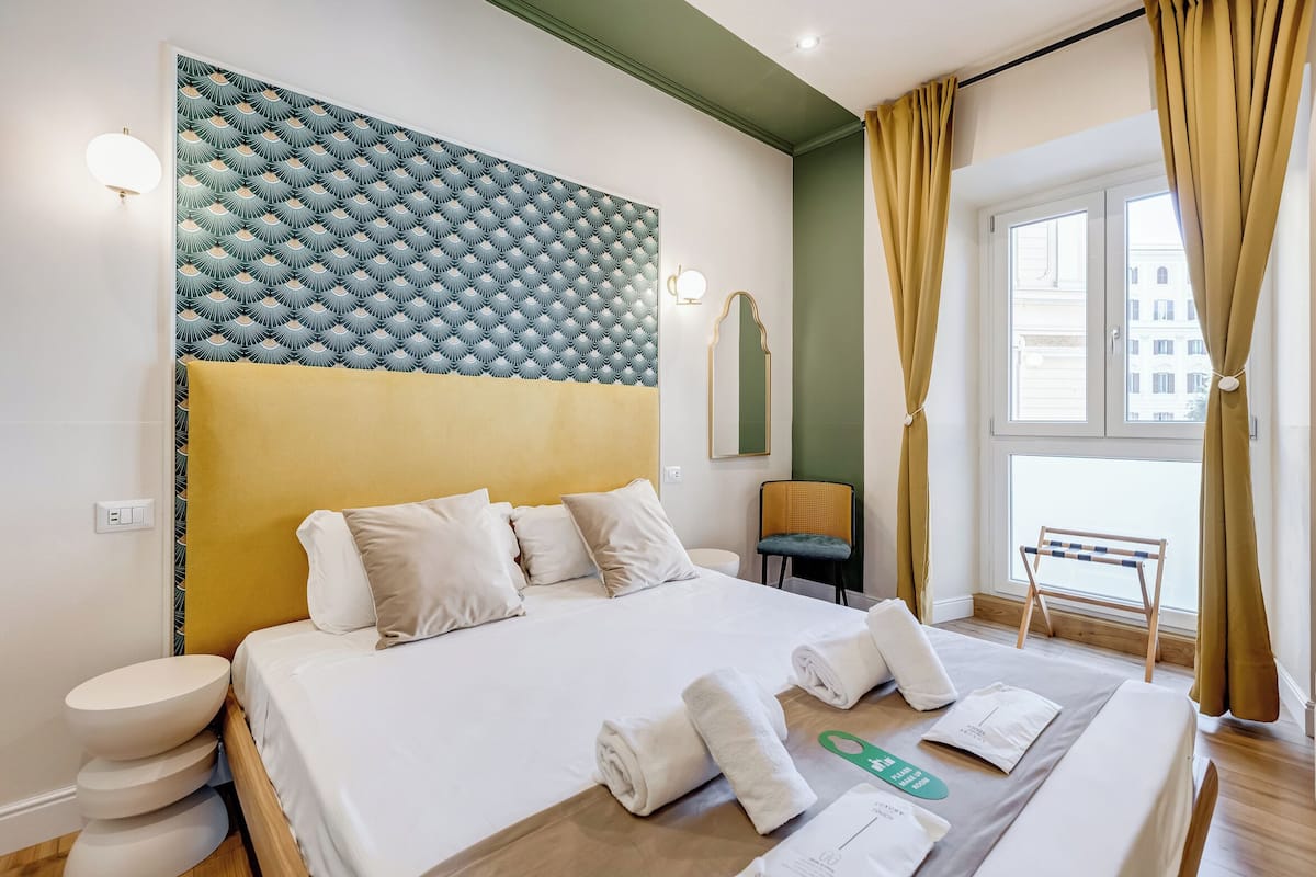 A hotel room with a large bed, a headboard with a geometric pattern, a bedside table, a chair, and a window with curtains.