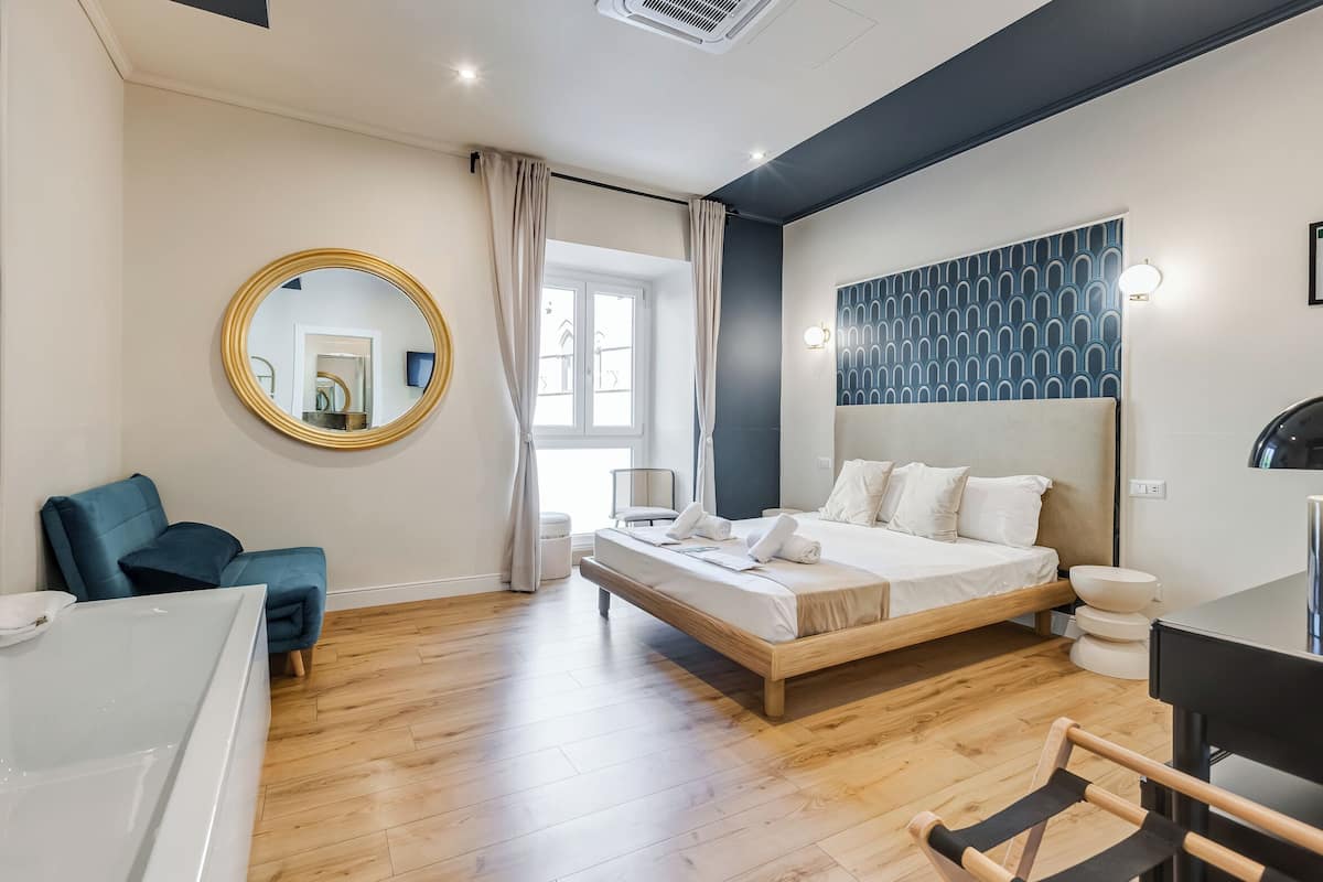 A modern bedroom with a large bed, a round mirror, a blue chair, and a white bathtub.