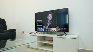 A modern living room with a flat-screen TV displaying a live stream, a glass coffee table, a black sofa, and a white entertainment unit with a book and remote controls. - Hawana Premium Hostel (Dubai)