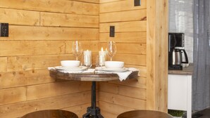 Un interior de madera con una mesa redonda dispuesta para dos personas, dos taburetes y una zona de cocina con cafetera.