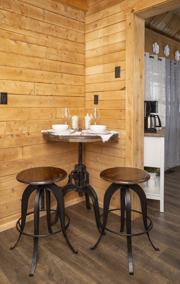 A wooden interior with a round table set for two, two stools, and a kitchen area with a coffee maker.