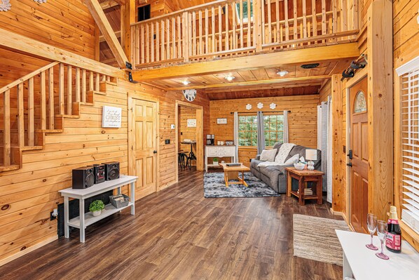 A wooden cabin interior with a staircase, a living room with a sofa and coffee table, and a dining area with a table and chairs.