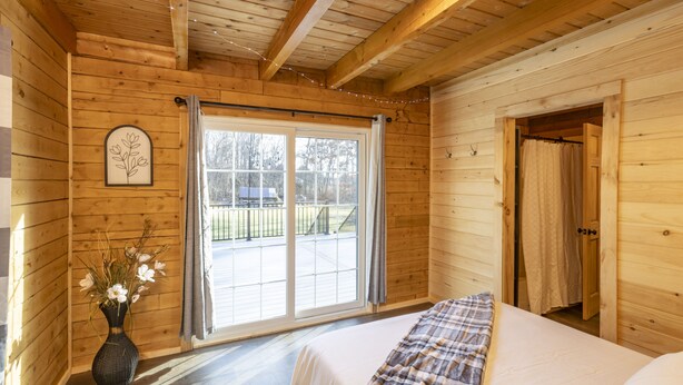 Una habitación de cabaña de madera con una cama, una ventana con cortinas, un jarrón con flores y una puerta que da a otra habitación.