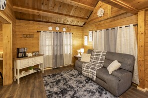 Ein gemütliches Zimmer mit Holzwänden, einem grauen Sofa, einem kleinen weißen Tisch mit einer Kaffeemaschine und einem flauschigen Teppich.