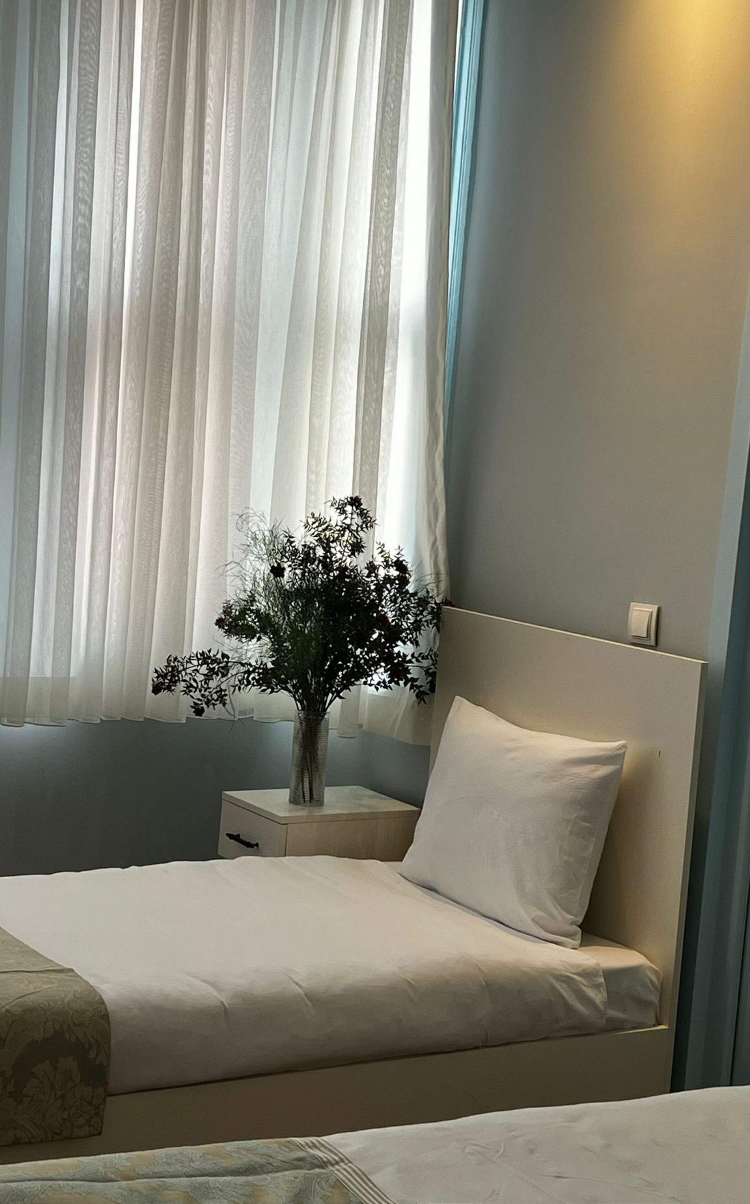 Una cama bien tendida con sábanas blancas, una mesita de noche con un florero y una ventana con cortinas translúcidas.