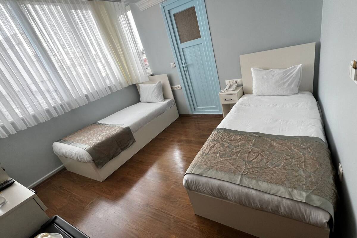 A bedroom with two beds, a bedside table, a nightstand, a window with sheer curtains, and a door.