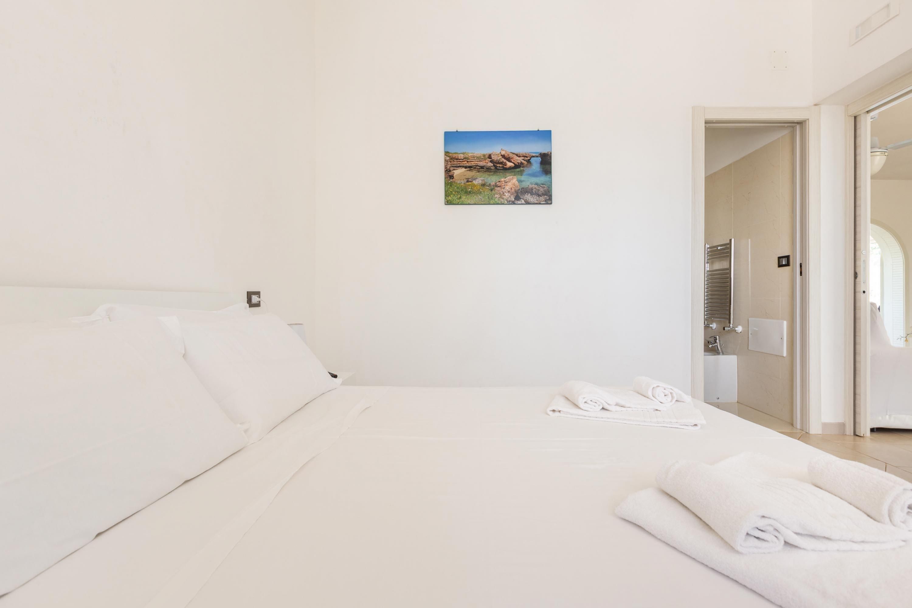 A hotel room with a bed, white linens, a painting of a coastal landscape, and a bathroom visible through an open door.