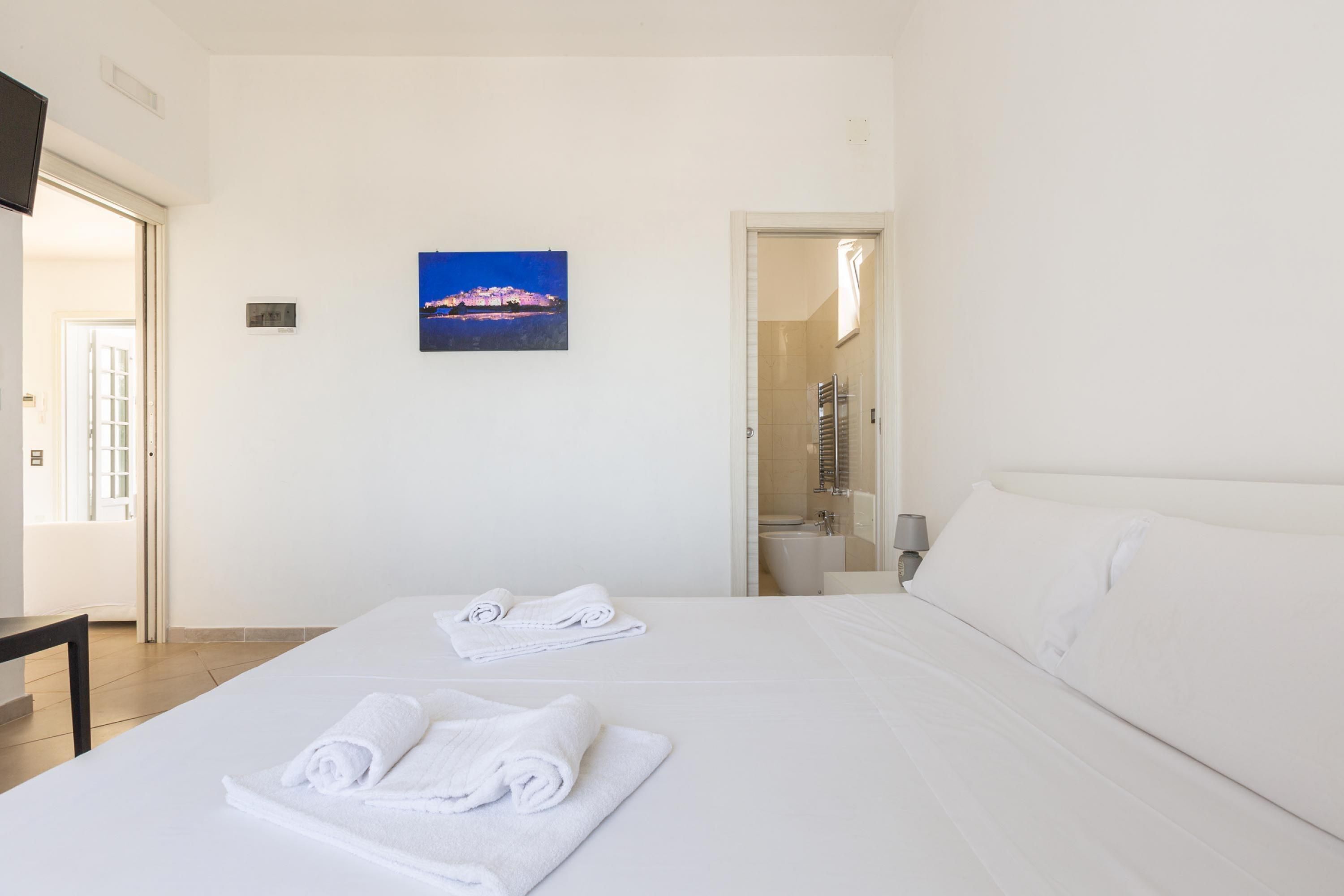 A hotel room with two beds, white linens, a painting of a landscape, a bathroom visible through an open door, and a flat-screen TV mounted on the wall.
