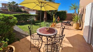 Een terras met een ronde tafel en stoelen, een gele parasol en potplanten.